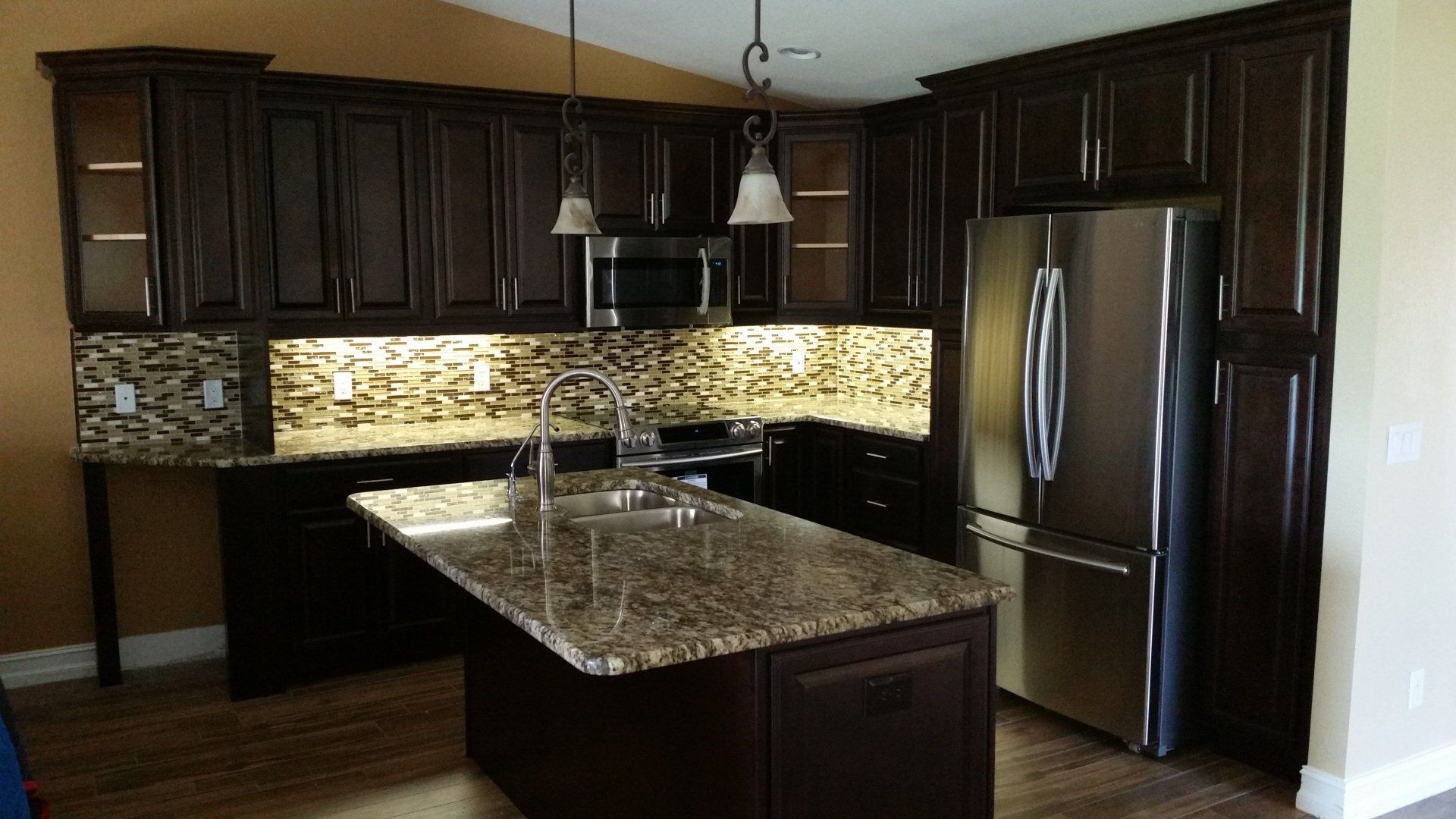 Dark brown kitchen with granite island, stainless steel appliances, and mosaic tile backsplash.