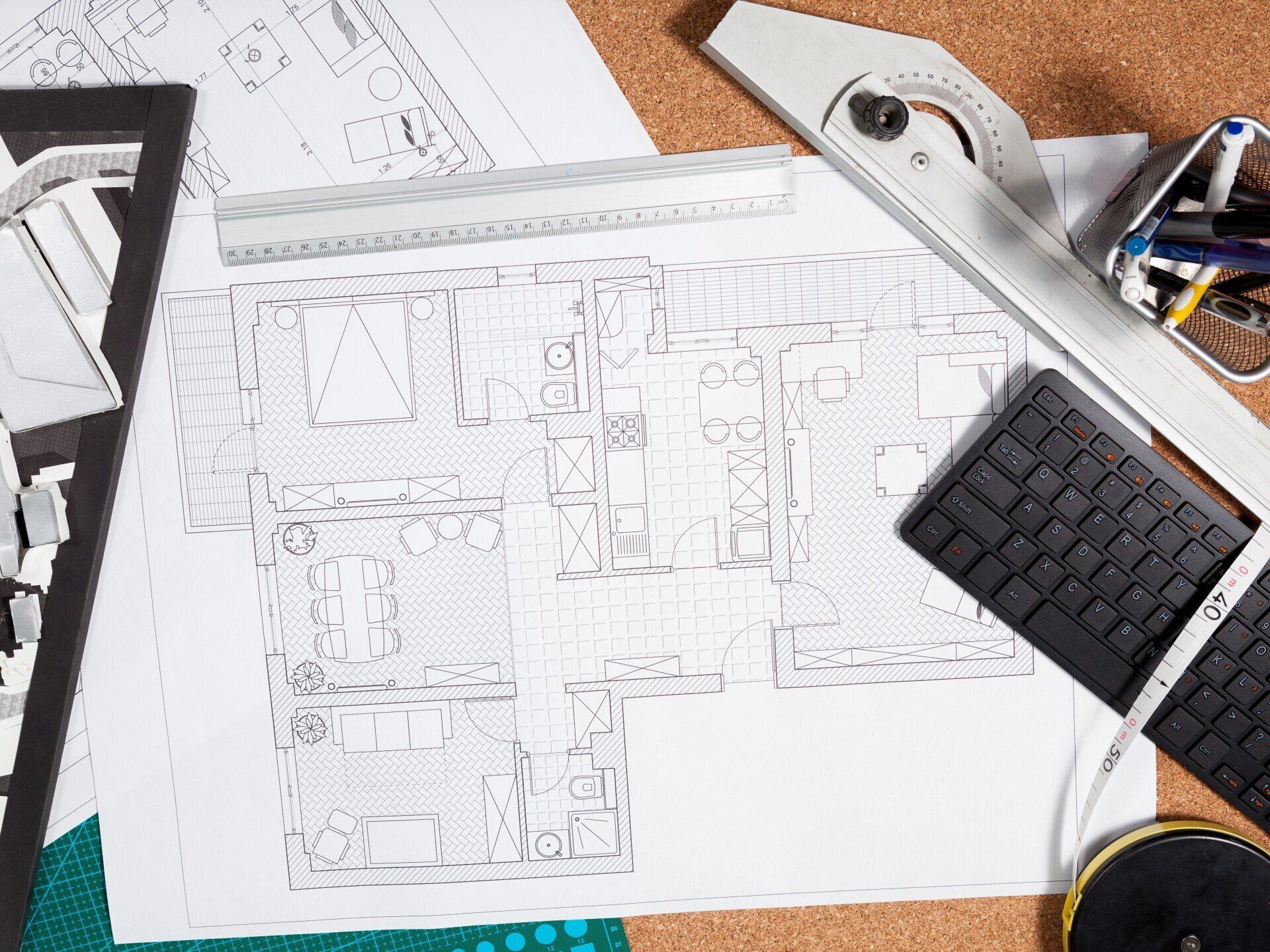 Architect's workspace: blueprints, ruler, protractor, keyboard, pens, and a model on a corkboard.
