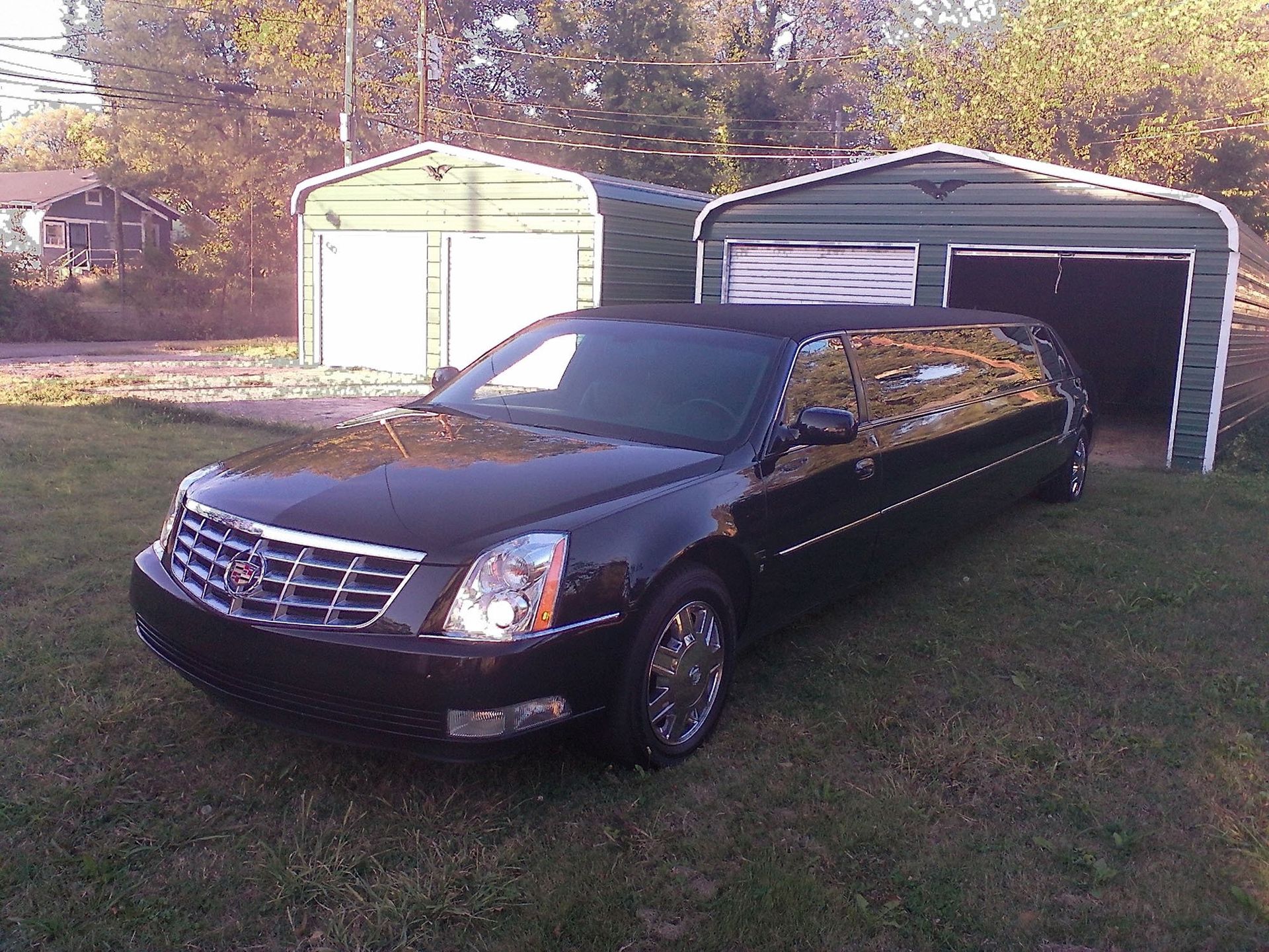 A black stretch limousine parked on a grassy lawn in front of two green metal storage garages.
