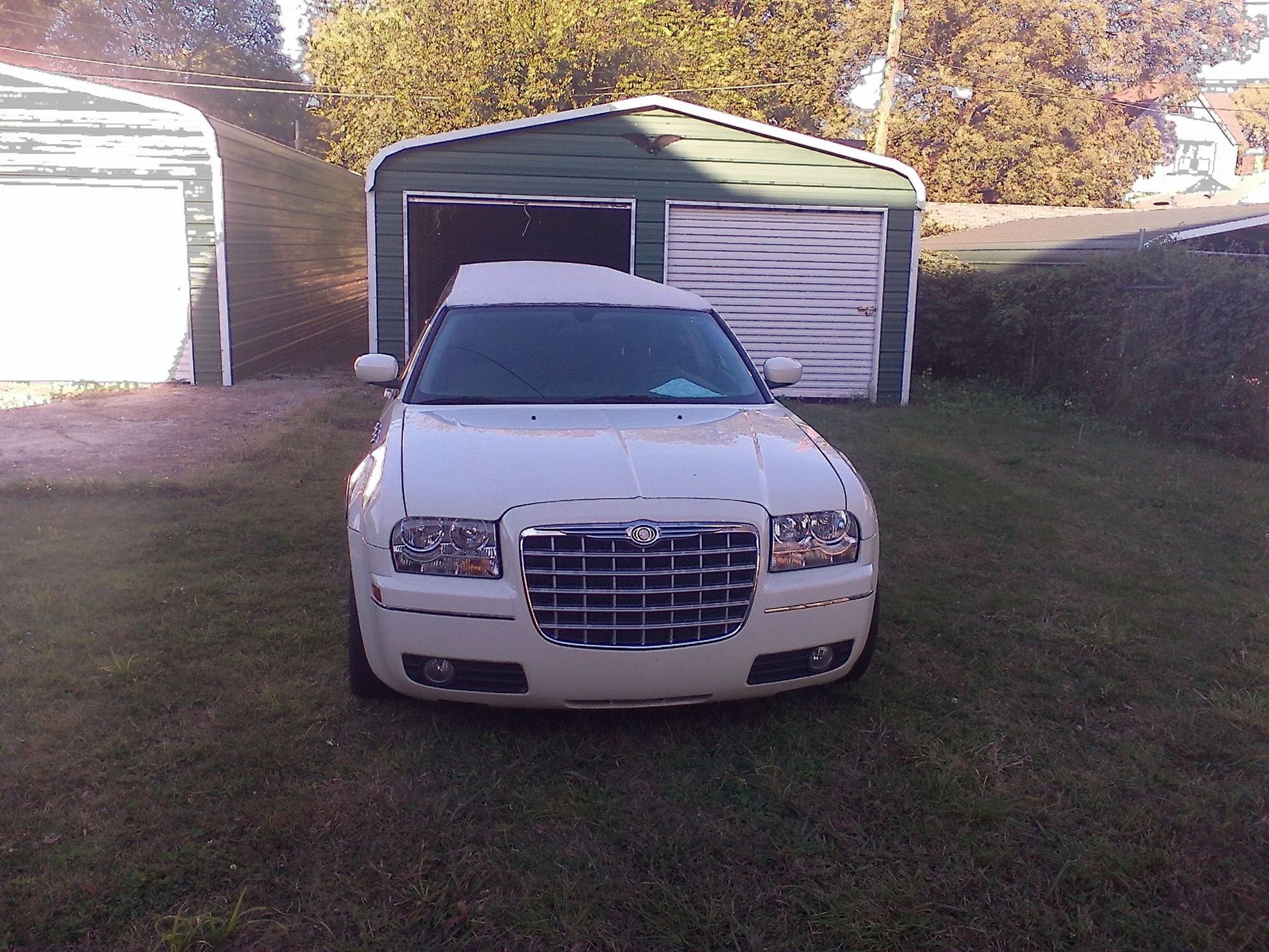 A white Chrysler 300 hearse parked on a grassy lawn in front of two metal storage sheds.