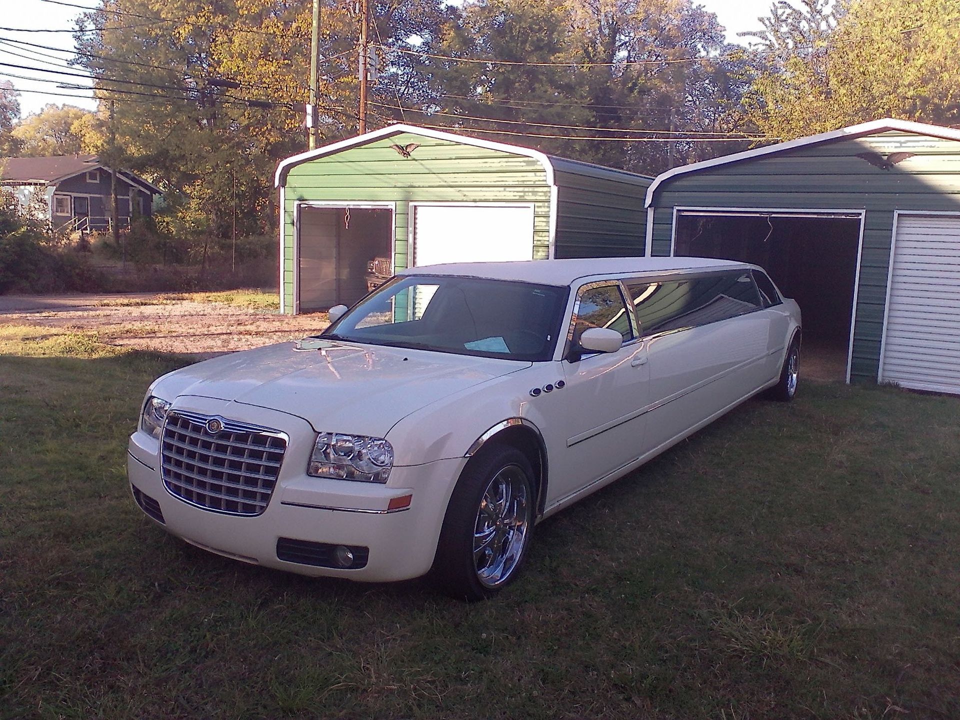 A white Chrysler 300 stretch limousine parked on a grassy lot in front of two green metal storage garages.