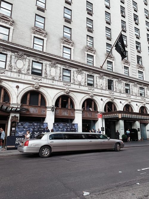 A silver limousine parked in front of a grand stone building with arched entrances and many windows.