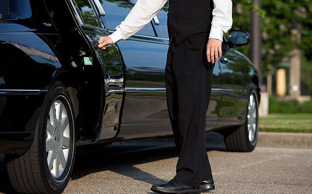 A chauffeur in a black suit holds open the door of a black limousine parked outdoors.