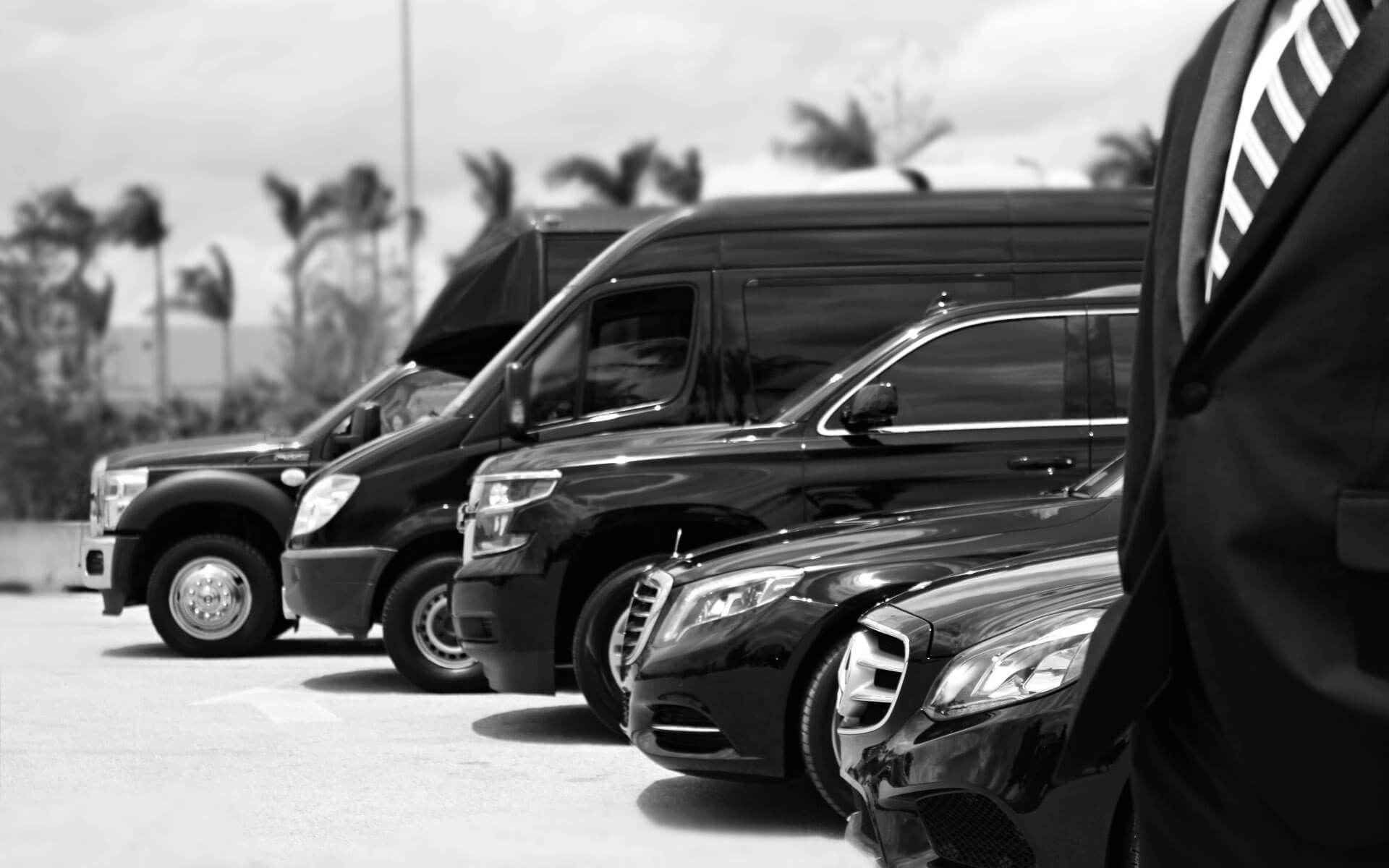 A lineup of parked black luxury vehicles and a van, with a person in a suit partially visible in the foreground.