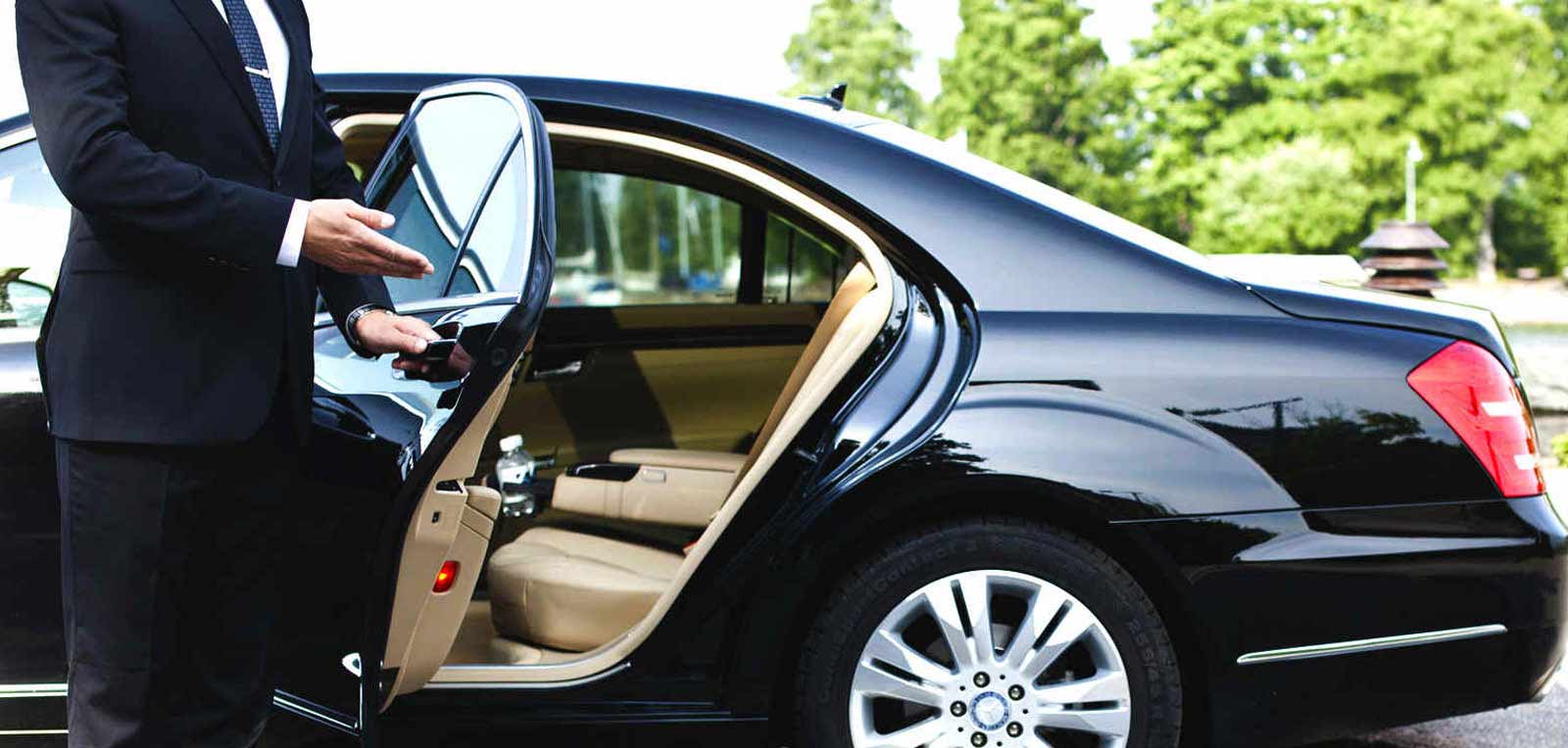 A professional driver in a suit holds open the door of a black luxury car to welcome a passenger.