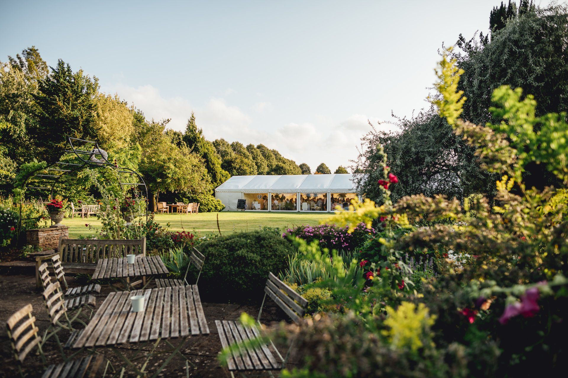 A large white tent is sitting in the middle of a garden surrounded by trees.