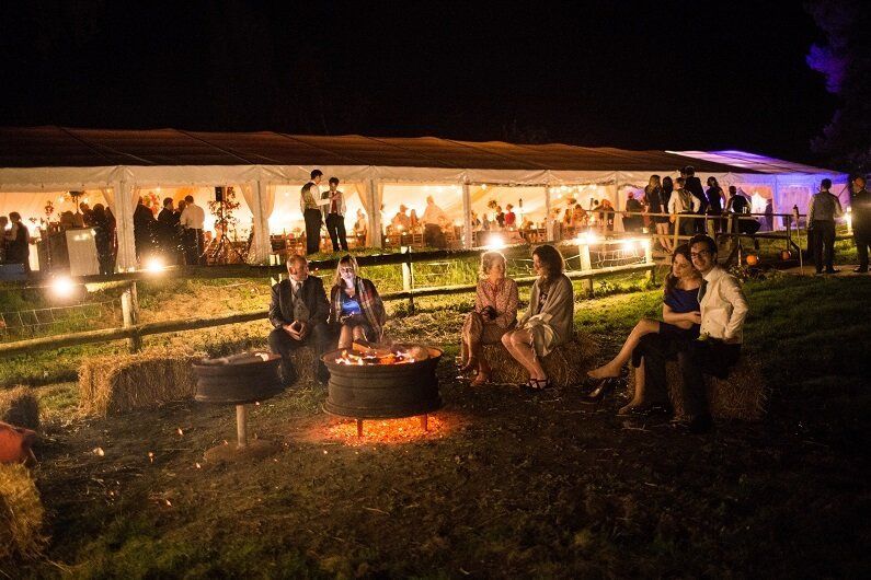 A group of people are sitting around a fire pit at night outside a wedding marquee on an Autumn evening
