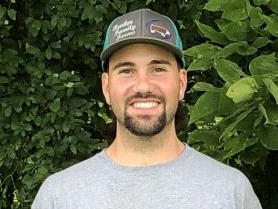 Smiling man in a gray shirt and dark cap standing in front of leafy greenery