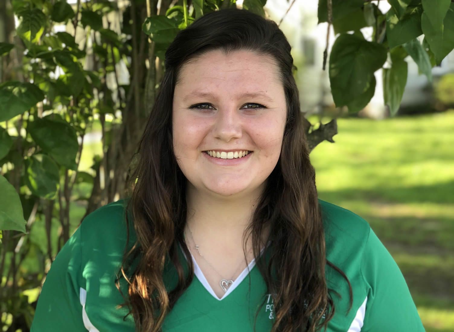 Smiling person in a green shirt standing outdoors among leafy trees.