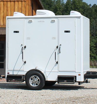White portable restroom trailer with double doors and small windows, parked on gravel outdoors