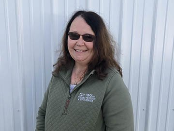 Smiling person in sunglasses and green jacket standing in front of a white corrugated wall