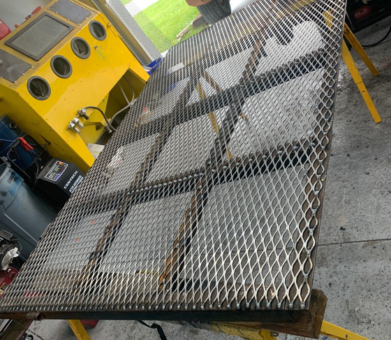 Expanded metal sheet on a steel frame, supported by yellow sawhorses. A yellow sandblasting cabinet is in the background.