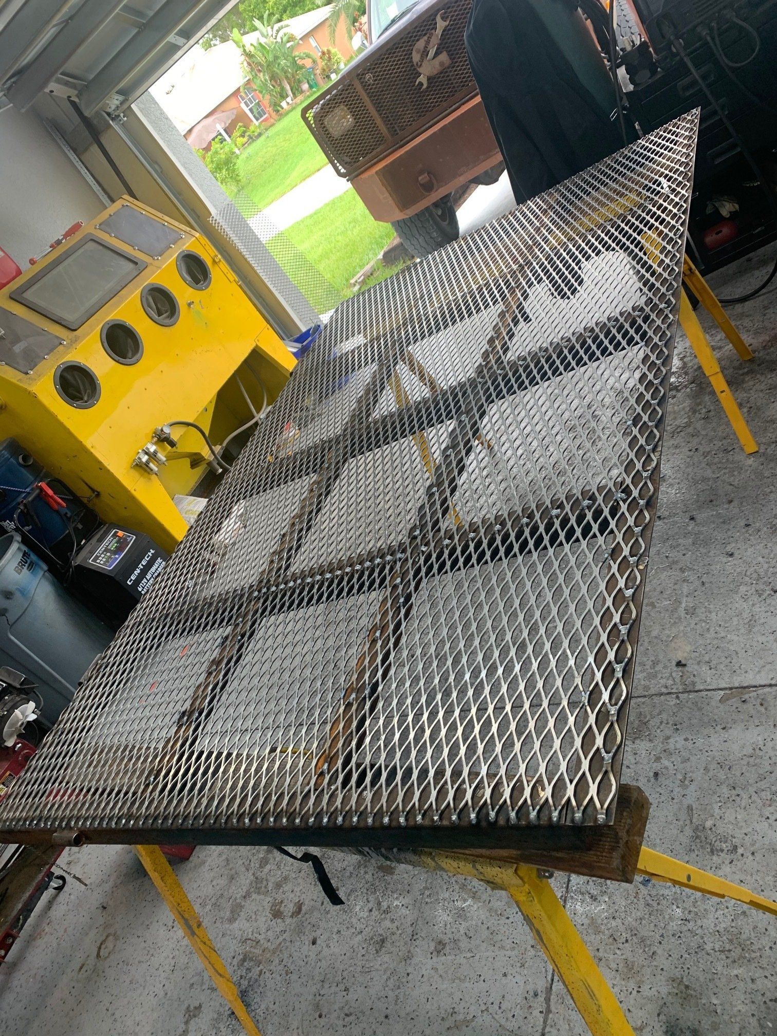 Large metal grate with welded supports on yellow sawhorses, inside a garage.