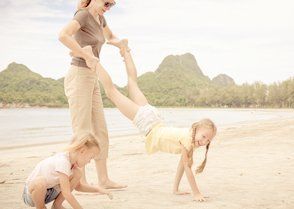 Een vrouw houdt de benen van een meisje vast terwijl het meisje een handstand maakt op het strand. Een ander meisje speelt in de buurt.