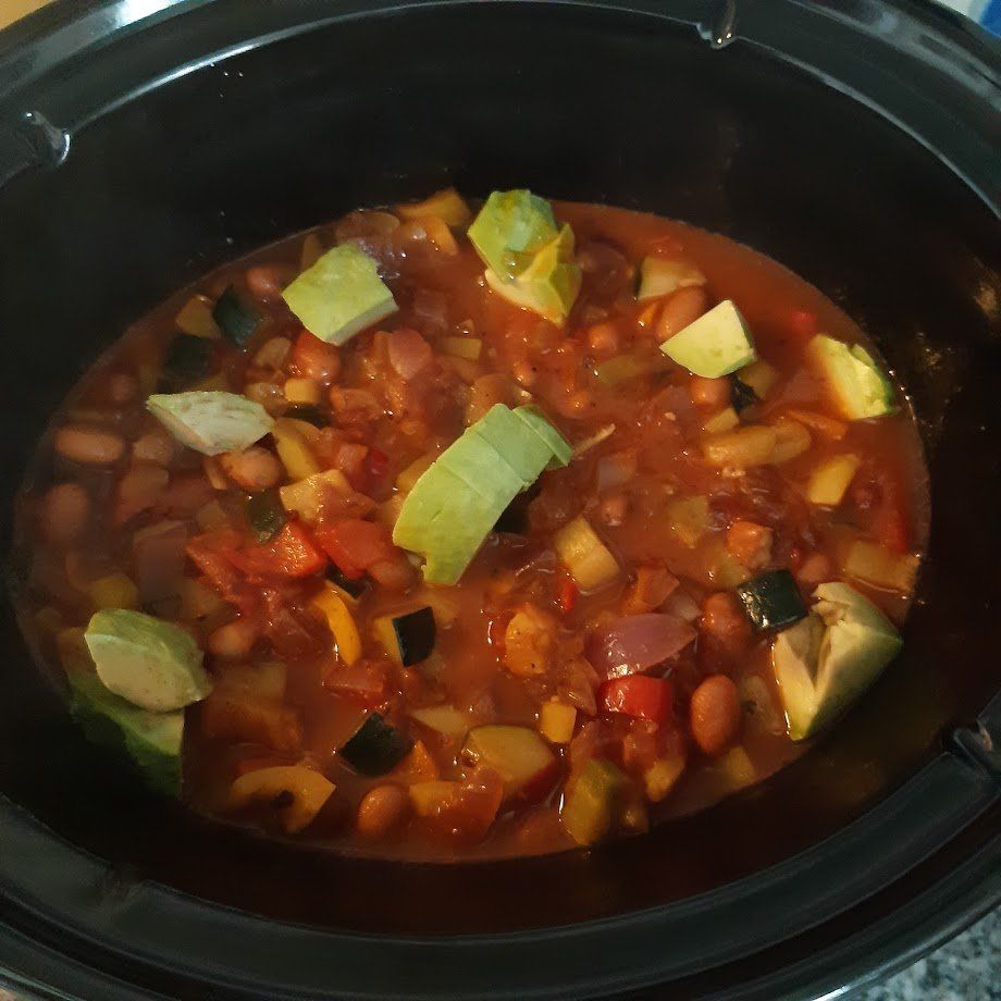 Chili con carne uit een zwarte slowcooker, met stukjes avocado, groenten en bonen.