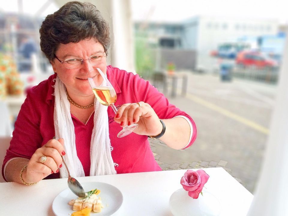 Vrouw ruikt aan champagne en proeft eten aan een tafeltje buiten; witte sjaal, roze blouse.