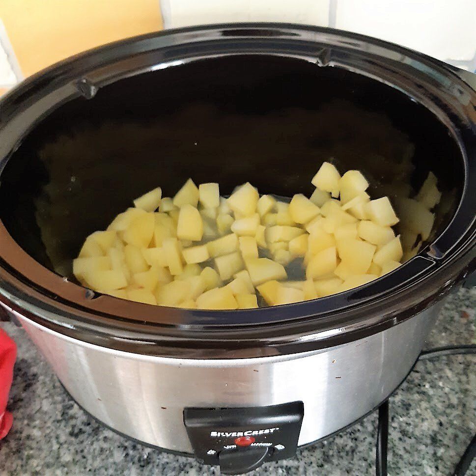 In blokjes gesneden aardappelen in een zwarte slowcooker, die op een grijs aanrecht staat.