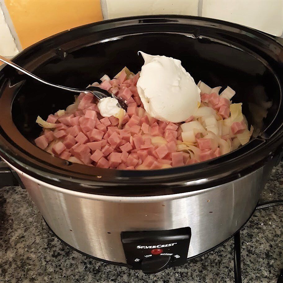 Een slowcooker gevuld met in blokjes gesneden ham, uien en een schepje witte room.