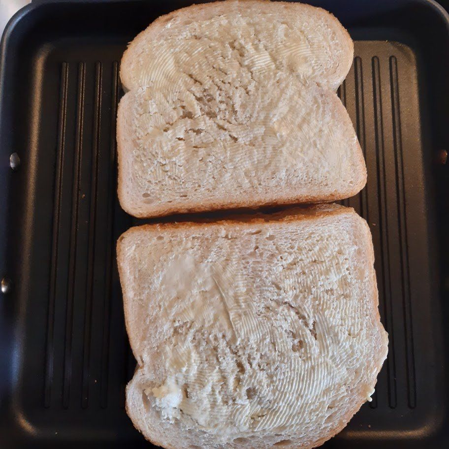 Twee sneetjes brood, met de boterkant naar beneden, in een zwarte grillpan.