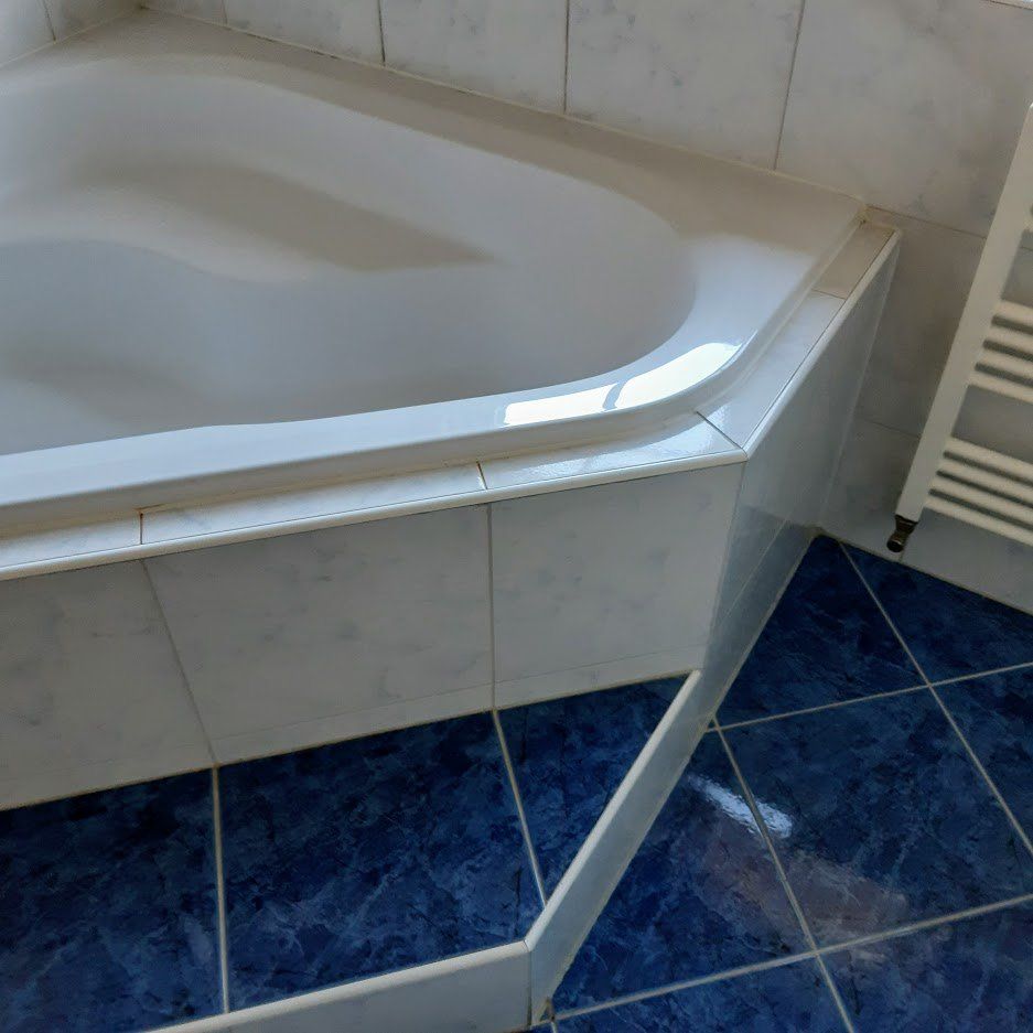 Witte badkuip met blauwe tegels rondom en op de vloer, in een hoek van de badkamer.