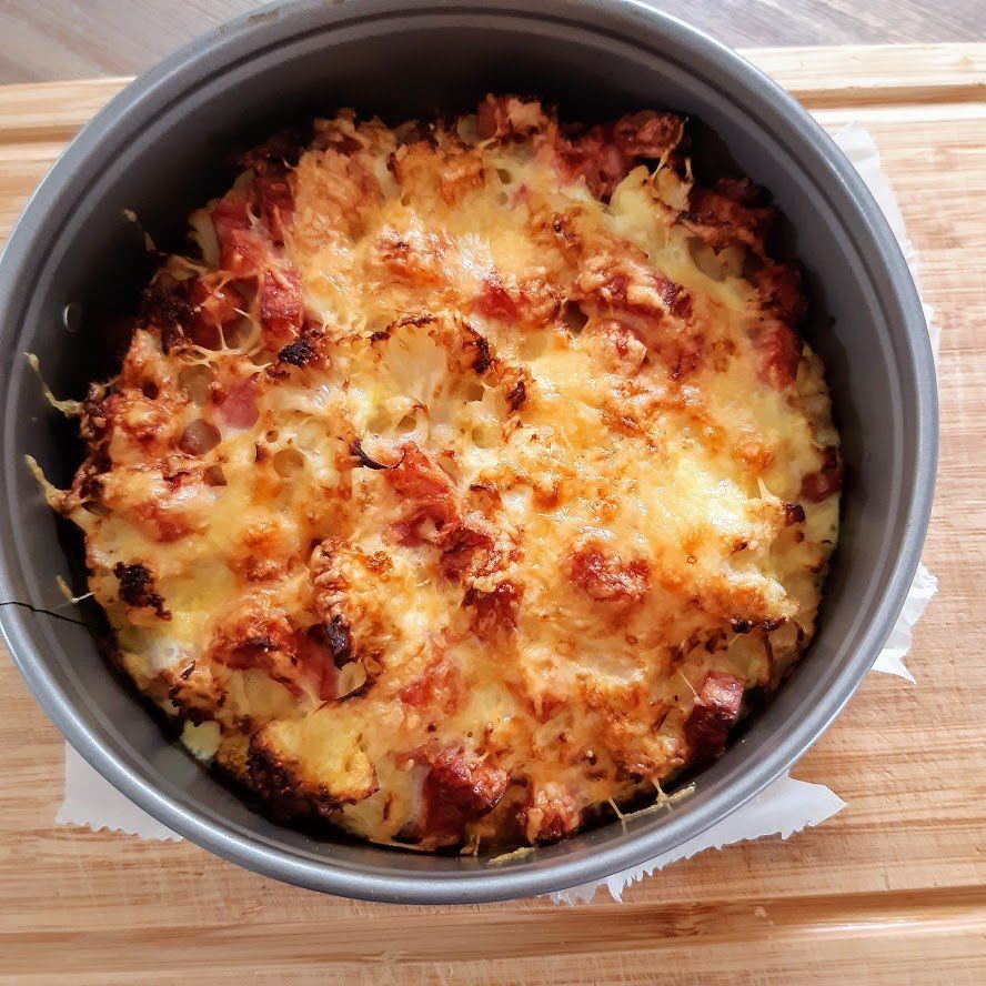 Ovenschotel in een metalen pan met gesmolten kaas, spek en bloemkool op een houten plank.