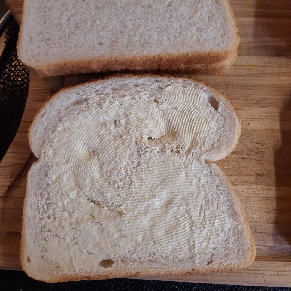 Een snee brood, beboterd, op een houten snijplank, de tweede snee erop.