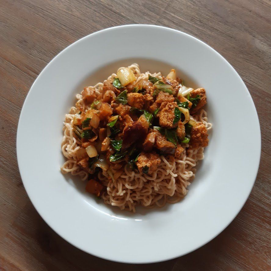 Een kom noedels met daarop een mengsel van tofu, ui en kruiden.