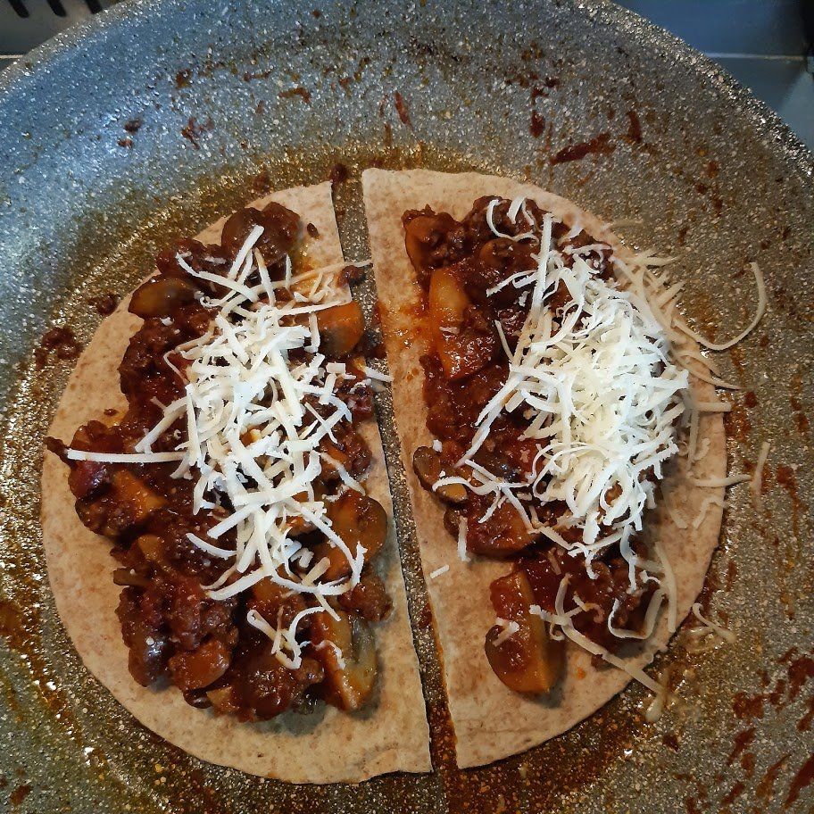 Twee quesadilla's gevuld met een hartige vulling en bedekt met geraspte kaas, gebakken in een pan.