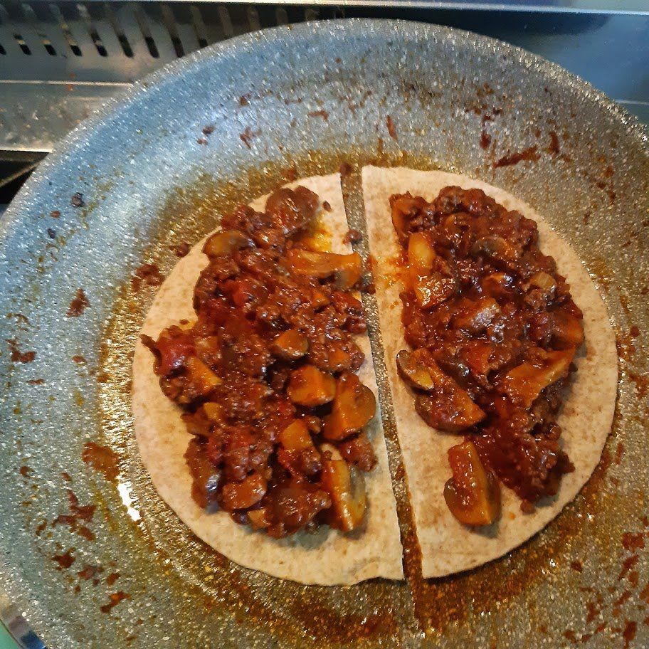 Een doormidden gesneden tortilla, belegd met champignons en rode saus, wordt in een pan op het fornuis gebakken.