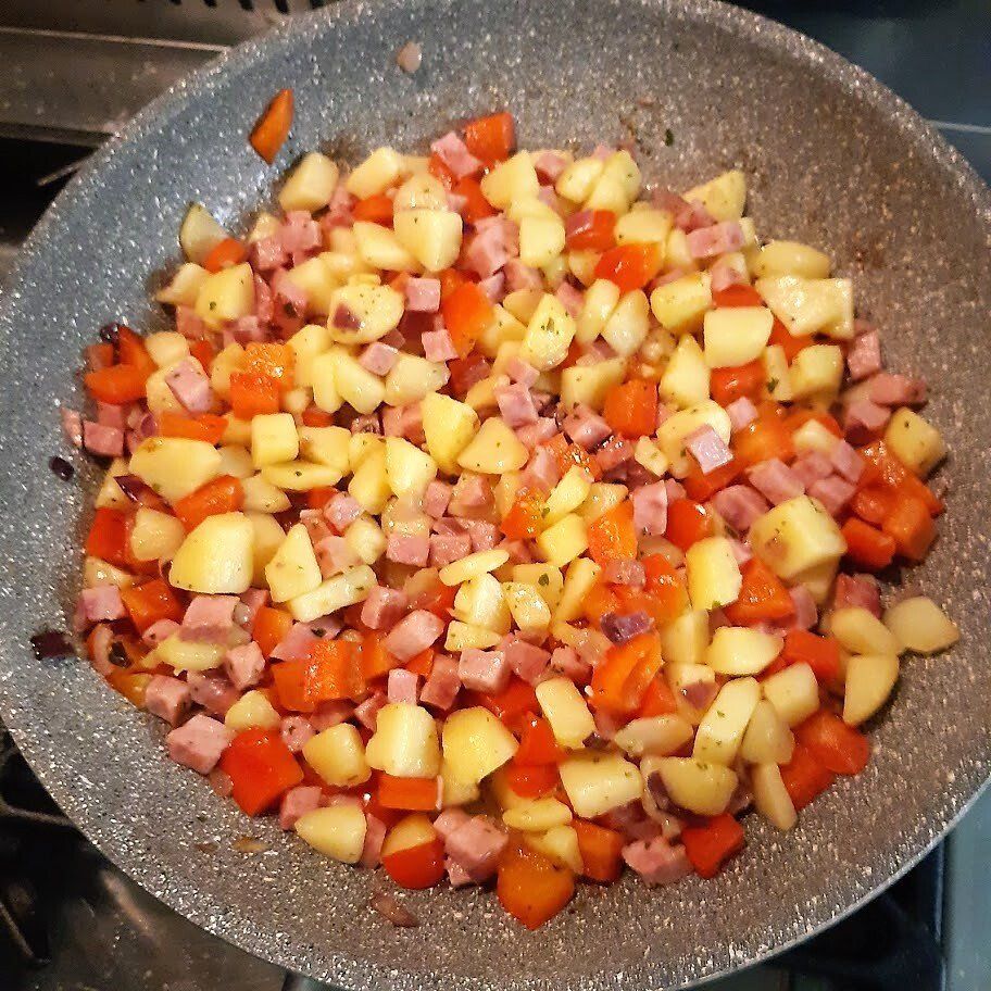 In een grijze pan op het fornuis worden blokjes aardappel, ham en rode paprika gebakken.