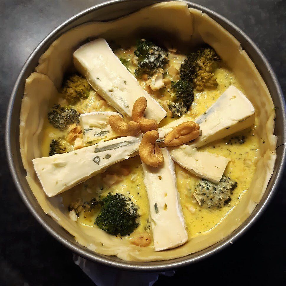 Een ongebakken quiche in een metalen bakvorm met broccoli, kaasblokjes en cashewnoten.