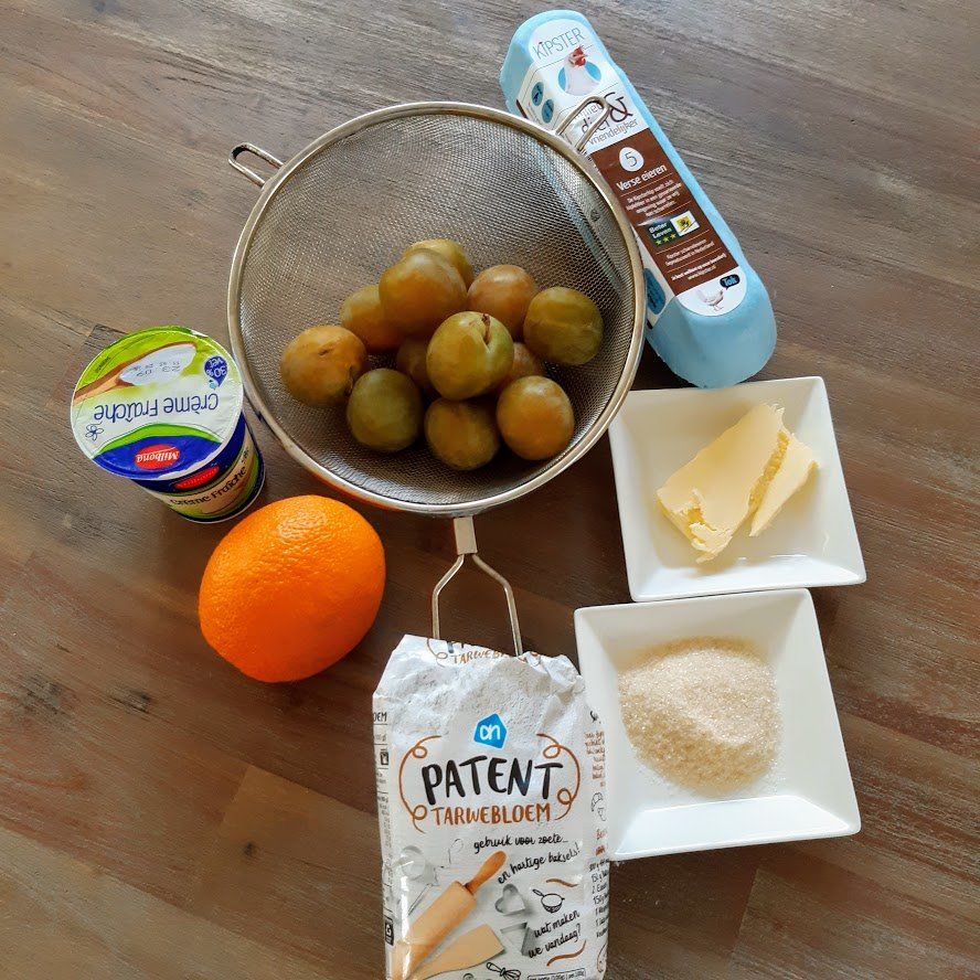 Ingrediënten voor een recept: pruimen in een zeef, bloem, suiker, boter, yoghurt, sinaasappel en een fles vloeistof.