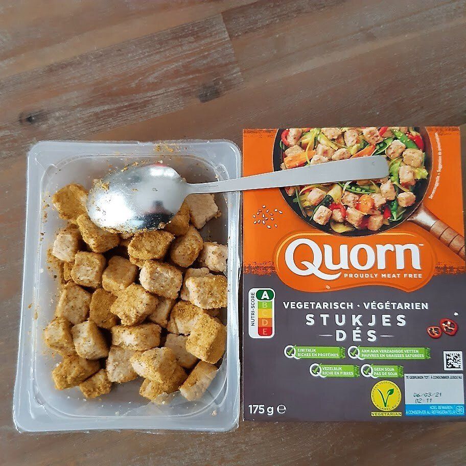 Vegetarische Quorn-stukjes in een bakje met lepel, naast de productdoos op een houten ondergrond.
