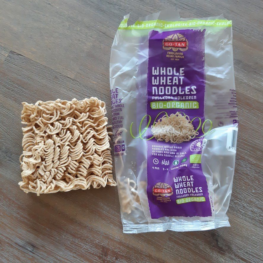 Een pak droge volkoren noedels en een blok ongekookte noedels op een houten ondergrond.