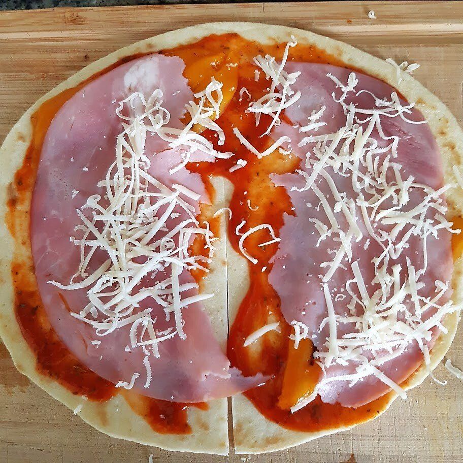 Pizzabodem met rode saus, plakjes ham en geraspte kaas op een houten ondergrond.