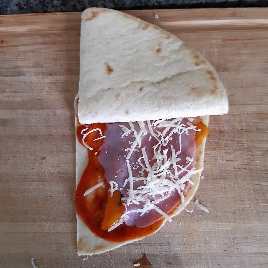 Een halfgevouwen tortilla met tomatensaus, ham, kaas en een plakje gele paprika op een houten plank.