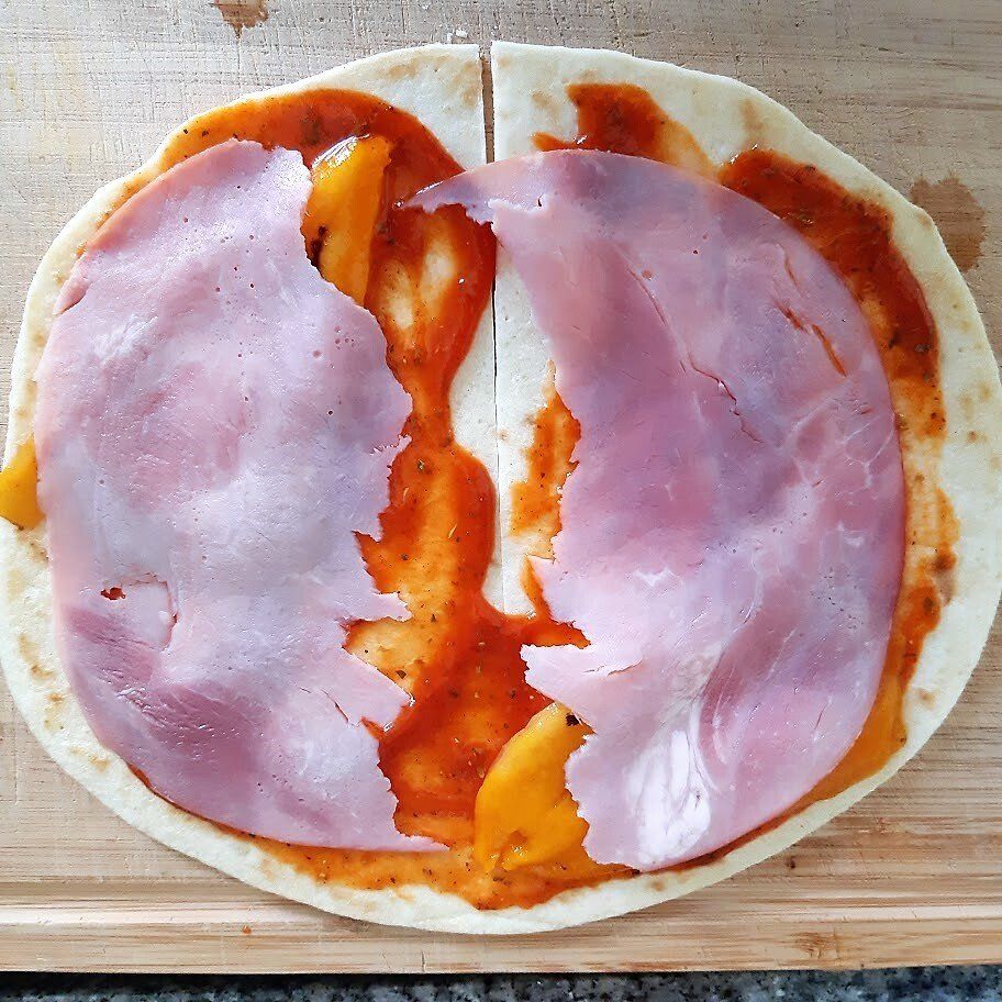 Een tortilla met saus, ham en kaas, klaar om te worden gevouwen en gebakken, op een houten oppervlak.