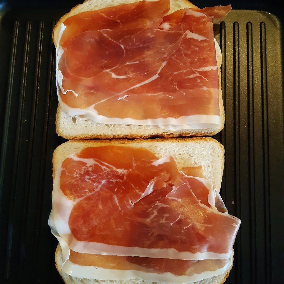 Twee sneetjes brood belegd met boterhammen en prosciutto, op een zwarte grill.