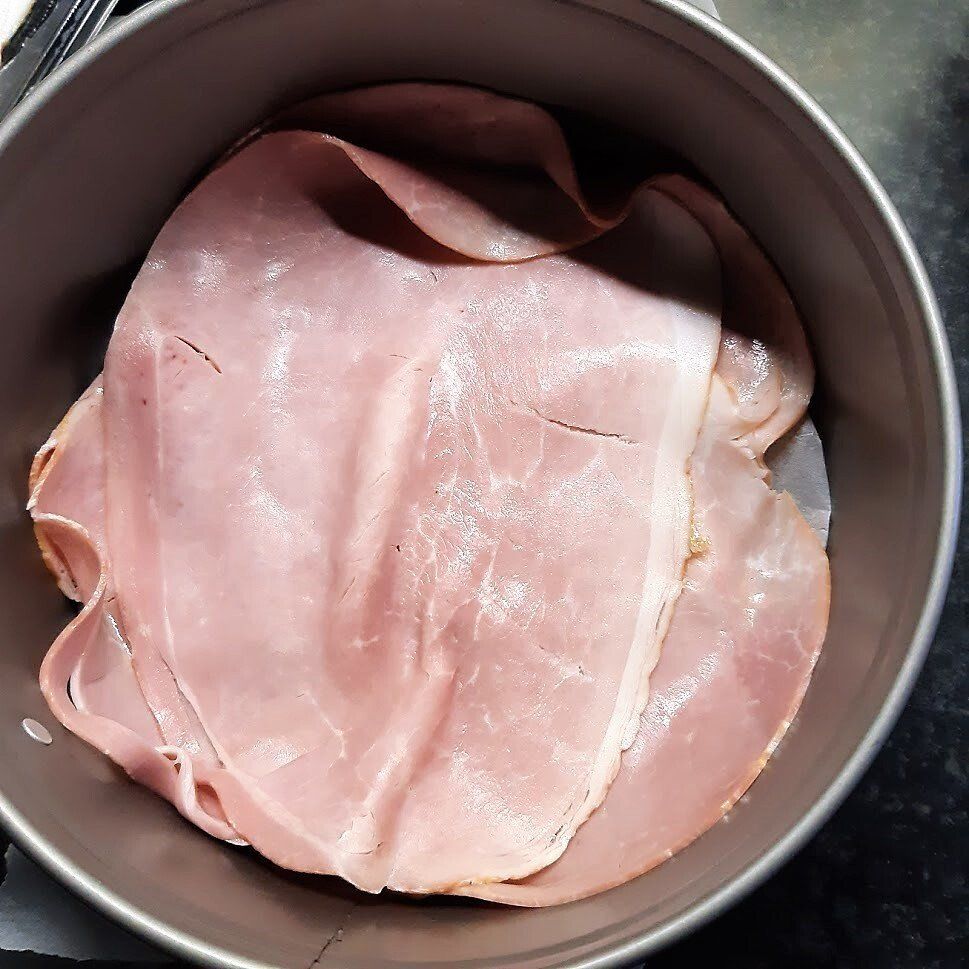 Plakjes roze ham in lagen in een ronde metalen ovenschaal.