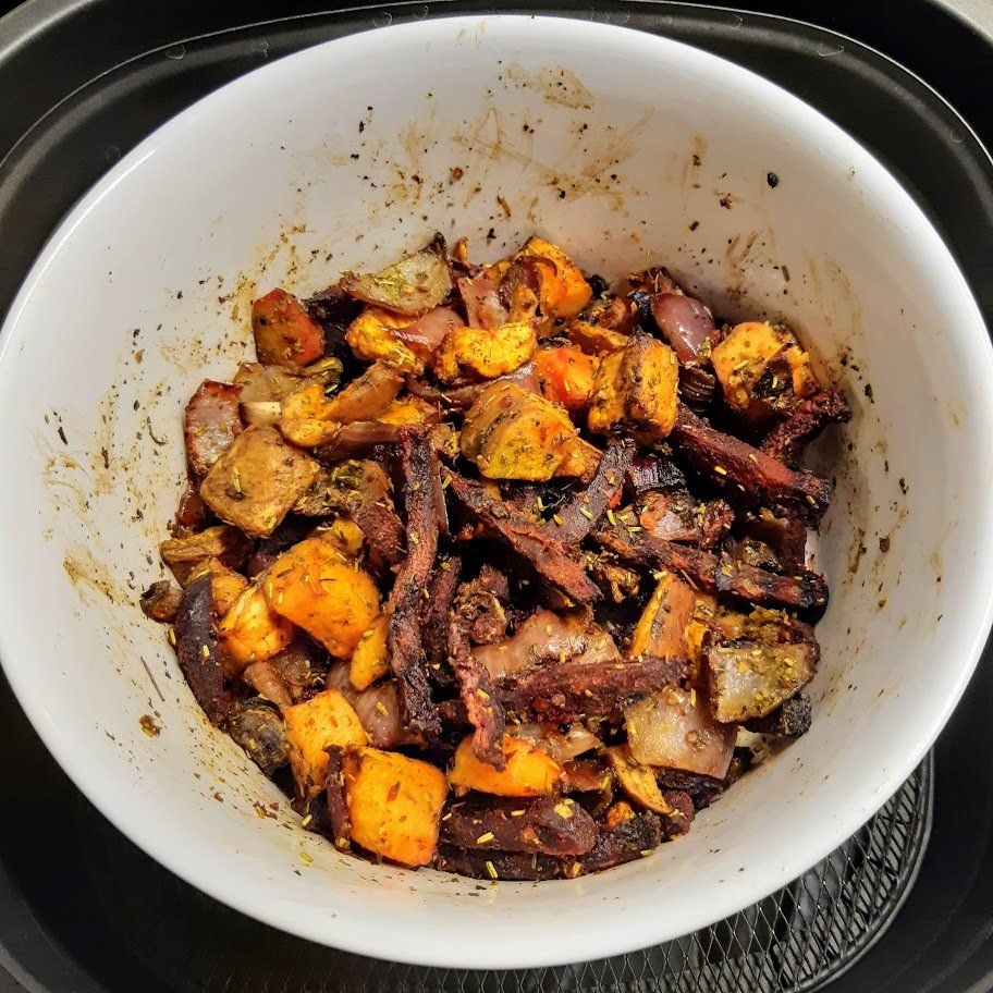 Geroosterde groenten in een witte kom in een airfryer. Oranje, paarse en bruine stukjes met kruiden.