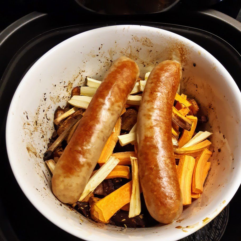 Twee worstjes en groenten in een witte kom in een zwarte airfryer.