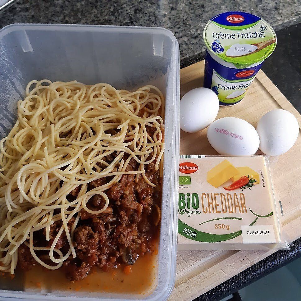 Pasta met vleessaus in een bakje, samen met eieren, kaas en crème fraîche op een snijplank.
