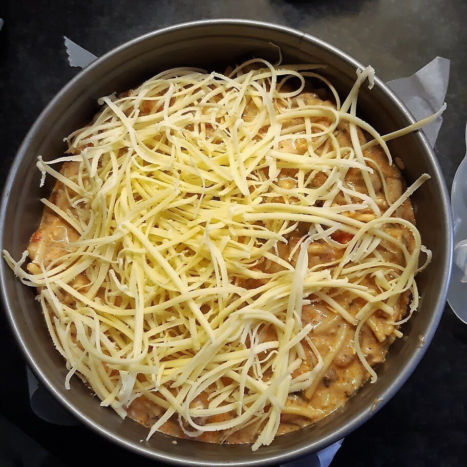 Ovenschotel in een ronde pan, bedekt met geraspte gele kaas. De romige saus is eronder zichtbaar.