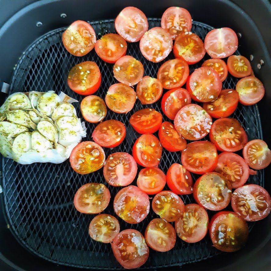 Een mandje voor de airfryer met gehalveerde tomaten en een bol knoflook, gekruid en klaar om te koken.