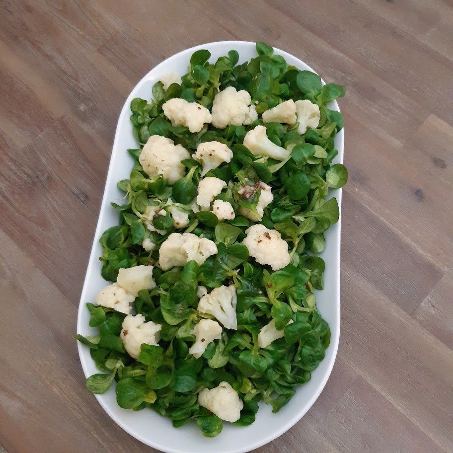 Een ovale witte schaal met groene bladsalade en bloemkoolroosjes op een houten tafel.