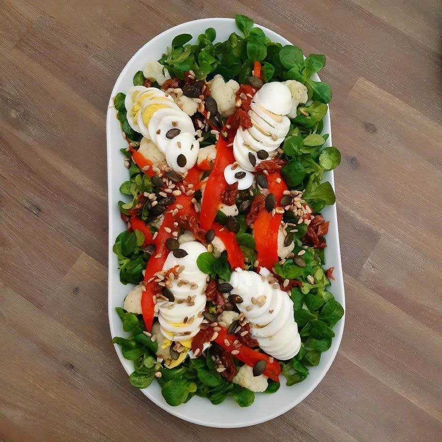 Ovale schaal met salade: groene bladgroenten, plakjes mozzarella, rode paprika, zaden en plakjes ei.