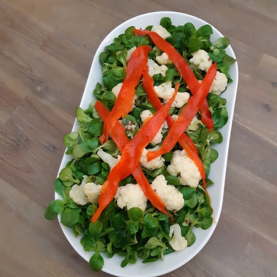 Salade in een witte schaal: groene bladeren, bloemkoolroosjes, reepjes rode paprika, op een houten tafel.