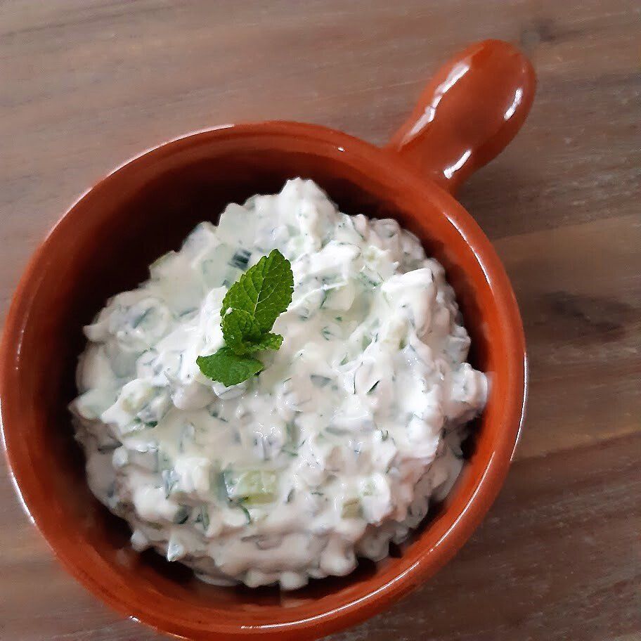 Serveer tzatziki in een klein bruin kommetje, gegarneerd met een takje munt, op een houten ondergrond.