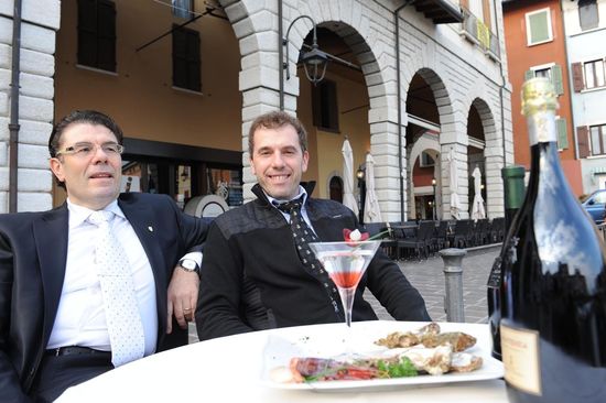 2 uomini seduti dietro un tavolo circolare bianco con cibo, bicchiere e bottiglia