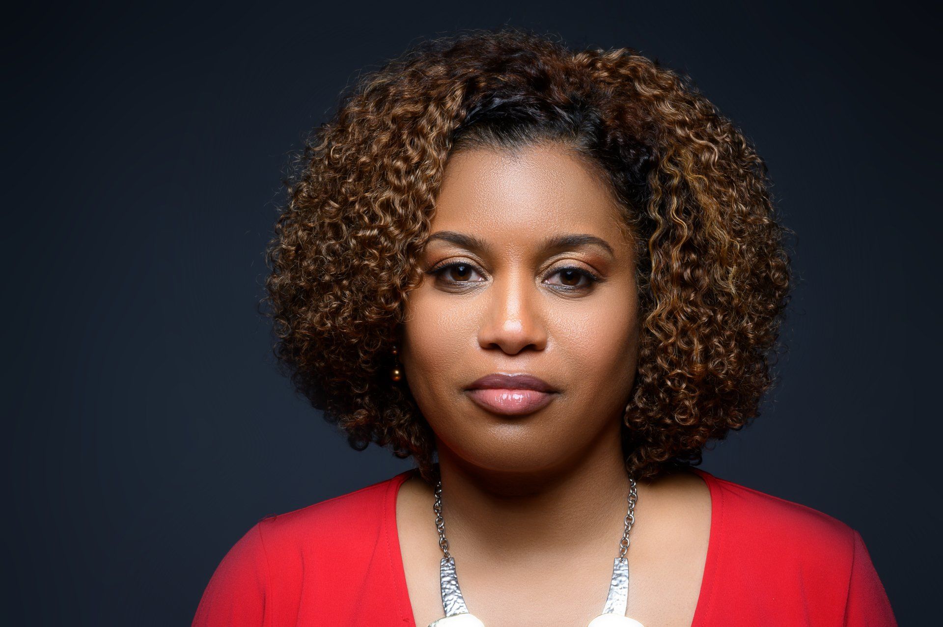 A woman with curly hair is wearing a red shirt and a silver necklace.